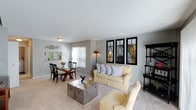 This two-floor residence offers a thoughtfully designed open-concept main level that seamlessly connects the living and dining areas. The living room is finished with plush wall-to-wall carpeting and features soft, neutral gray walls that lend a calm, versatile backdrop to the space. A comfortable sofa anchors the seating area, complemented by an accent armchair and a metallic trunk-style coffee table that adds an industrial touch. Decorative framed mirror panels with geometric detailing line one wall, enhancing the sense of depth and light in the room. A tall floor lamp provides warm ambient lighting beside the sofa. The adjoining dining area accommodates a glass-top table with cross-legged wooden chairs, ideal for everyday dining and entertaining. A window allows natural light to filter into the dining space, brightening the area. A dark wood display cabinet near the entry provides additional storage and visual interest. The kitchen is partially visible through a pass-through opening, suggesting a connected and practical layout. The overall ambiance is clean, bright, and move-in ready, offering a comfortable and cohesive living environment across both floors.