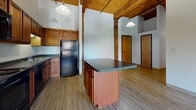 Located at 45 Willow St in Springfield, MA, this loft-style unit blends industrial character with comfortable living. The open-concept main area is immediately striking, featuring soaring ceilings clad in warm wood planking, supported by prominent exposed timber beams that run the length of the space. Original exposed brick walls add texture and historic charm, complementing the light hardwood-style flooring throughout the main living area.

The kitchen is well-appointed with rich cherry-toned cabinetry, dark granite-style countertops, and a full suite of black appliances, including a range, built-in microwave, dishwasher, and refrigerator. A central kitchen island with a matching countertop provides additional workspace and casual seating. Pendant lighting fixtures hang from the beamed ceiling, adding warmth to the cooking and dining area.

The bedroom features soft carpeting, generous wall space, and tall ceilings that carry the loft aesthetic throughout. Large arched windows along the exposed brick wall allow natural light to fill the space while offering views of the surrounding neighborhood. A full bathroom with a tub/shower combination and wood-toned cabinetry completes the layout. The overall ambiance is one of urban industrial elegance, ideal for those who appreciate character-rich architectural details.