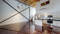 This two-story loft showcases a striking industrial aesthetic rooted in its warehouse origins. The main level immediately impresses with its soaring ceiling height, exposed wood-decked steel roof trusses, and a large-blade ceiling fan that underscores the expansive volume of the space. Polished, acid-stained concrete floors reflect light across the open-plan living and kitchen area, while dramatic black steel X-brace wall detailing serves as a bold architectural focal point. Track lighting illuminates the space throughout, and windows along the main level allow natural light to filter in.

The kitchen is well-appointed with white shaker cabinetry, dark countertops, a mosaic tile backsplash, stainless steel appliances, and a central island. A staircase with wood treads leads to the upper level, which features wood-look plank flooring, an exposed truss ceiling, and a spacious bedroom with double doors opening to a walk-in closet. An interior window overlooks the dramatic main-level volume below.

The bathroom presents upscale finishes including large-format marble-style tile in the shower, a vessel sink atop dark counters, an oval mirror, and polished concrete flooring — cohesively tying together the industrial and contemporary design elements found throughout the space.