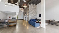 Located at 1000 Diamond St in Philadelphia's 19122 zip code, this contemporary lobby space offers a striking first impression with its dramatic double-height ceilings and expansive floor-to-ceiling glass entry doors that flood the interior with natural light. The polished concrete floors extend throughout, complementing the industrial-chic aesthetic defined by exposed ductwork and boldly designed pendant lighting fixtures.

A defining architectural feature is the full-height stacked stone accent wall, which rises to the ceiling and anchors the main lounge area. This seating area is furnished with a blue sectional sofa, a coffee table, and a wall-mounted flat-screen TV, creating a comfortable and welcoming atmosphere. A second decorative stone accent wall near the elevator corridor adds warmth and texture throughout the space.

The lobby provides access to two elevators, a stairwell, and adjacent tenant spaces, including a unit marked as Night Owls Fast Food. A secondary lounge area near the entrance features a living green wall installation and additional console tables with potted plants, introducing a natural element that softens the industrial design. The space balances modern commercial design with inviting, carefully curated details, making it a functional and visually engaging communal hub for the building's occupants and visitors.