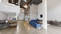 Located at 1000 Diamond St in Philadelphia's 19122 zip code, this contemporary lobby space offers a striking first impression with its dramatic double-height ceilings and expansive floor-to-ceiling glass entry doors that flood the interior with natural light. The polished concrete floors extend throughout, complementing the industrial-chic aesthetic defined by exposed ductwork and boldly designed pendant lighting fixtures.

A defining architectural feature is the full-height stacked stone accent wall, which rises to the ceiling and anchors the main lounge area. This seating area is furnished with a blue sectional sofa, a coffee table, and a wall-mounted flat-screen TV, creating a comfortable and welcoming atmosphere. A second decorative stone accent wall near the elevator corridor adds warmth and texture throughout the space.

The lobby provides access to two elevators, a stairwell, and adjacent tenant spaces, including a unit marked as Night Owls Fast Food. A secondary lounge area near the entrance features a living green wall installation and additional console tables with potted plants, introducing a natural element that softens the industrial design. The space balances modern commercial design with inviting, carefully curated details, making it a functional and visually engaging communal hub for the building's occupants and visitors.