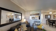 This single-floor residence offers a thoughtfully designed living space with a cohesive and contemporary aesthetic. The living room serves as the heart of the home, furnished with a plush sofa and patterned accent chairs accented by an array of decorative pillows in earthy tones of yellow, orange, and gray. A large, dark-framed wall mirror adds depth to the space, reflecting natural light and visually expanding the room. Abstract landscape artwork adorns the walls, contributing to a curated, gallery-like atmosphere. A textured popcorn ceiling and neutral gray walls provide a clean backdrop that complements the bold furnishings. A pass-through window connects the living area to the kitchen, offering a glimpse of cabinetry and appliances, and promoting an open, communicative flow between spaces. The kitchen features warm wood-toned cabinetry, hinting at a functional and well-equipped cooking area. A hallway visible in the background suggests additional rooms beyond, completing the layout of this single-level home. Overall, the space balances comfort and style with warm lighting, decorative accents, and a welcoming atmosphere suited for everyday living.