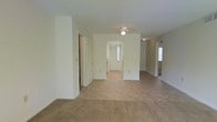 This single-story space features an open, flowing layout bathed in neutral cream and beige tones throughout. The main living area is generously sized, with light-colored carpet underfoot that transitions into tile flooring toward the central hallway, creating a natural visual distinction between spaces. A flush-mount ceiling light fixture illuminates the hallway, which serves as the connective corridor leading to additional rooms. Doorways branch off in multiple directions, suggesting a functional and well-organized floor plan. Windows at the far end of the hallway and to the right allow natural light to filter into the interior, brightening the otherwise understated palette. The walls are smooth and painted in a consistent off-white hue, contributing to the clean, uniform aesthetic. A second ceiling fixture is visible deeper in the space, indicating consistent overhead lighting throughout. The layout appears practical and straightforward, with clearly defined zones for living and utility areas. Overall, the space presents a simple, move-in-ready environment with a neutral design that offers flexibility for a variety of furnishing and décor styles.