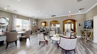 This single-story community clubhouse and leasing center is thoughtfully designed to provide both functional and welcoming spaces for residents and visitors. The main lounge area serves as the heart of the space, featuring wood-look flooring, crown molding, and recessed lighting throughout. Elegant arched doorways connect the various zones, lending an architectural continuity to the layout. Geometric-patterned accent chairs surround a round white coffee table, creating an inviting seating arrangement bathed in natural light from large windows dressed with patterned drapery.

A dedicated leasing office provides a private workspace with a curved desk, guest seating, and views into adjacent areas through decorative arched openings. A coffee bar station nearby offers convenience with white cabinetry, granite countertops, and bar seating beside bright windows.

The business center includes multiple workstations equipped with computers and a printer, accented by a bold teal feature wall and globe pendant lighting. A conference-style table with upholstered seating offers additional collaborative workspace beneath a gold orb chandelier.

A cozy lounge nook anchors one end of the space, featuring a tile-surround fireplace flanked by vibrant blue accent chairs and gold wall art, creating a warm and inviting retreat.