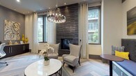 This contemporary amenity lounge is designed to deliver both comfort and functionality in an elegantly curated setting. The centerpiece of the main lounge area is a floor-to-ceiling dark tile fireplace wall, flanked by large windows dressed with patterned drapery that allow generous natural light and views of the surrounding outdoor courtyard. A statement chandelier with exposed Edison-style bulbs hangs overhead, adding warmth and sophistication to the space. Plush wingback chairs and a round marble-top table create an inviting seating arrangement ideal for relaxation or casual conversation. The herringbone-patterned carpet flooring runs throughout, lending a refined, textural quality underfoot. A secondary seating nook features a built-in upholstered bench with a bold patterned fabric, complemented by bistro-style side tables and a wall-mounted flat-screen television — perfect for more informal gatherings. Recessed lighting and curated artwork add further polish to the space. A dedicated business center alcove provides a curved built-in workstation with multiple desktop computers and printing capabilities, catering to productivity needs. The overall design palette of neutral tones with accent colors of navy and gold creates a cohesive, modern aesthetic throughout the lounge.