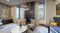 This contemporary amenity lounge is designed to deliver both comfort and functionality in an elegantly curated setting. The centerpiece of the main lounge area is a floor-to-ceiling dark tile fireplace wall, flanked by large windows dressed with patterned drapery that allow generous natural light and views of the surrounding outdoor courtyard. A statement chandelier with exposed Edison-style bulbs hangs overhead, adding warmth and sophistication to the space. Plush wingback chairs and a round marble-top table create an inviting seating arrangement ideal for relaxation or casual conversation. The herringbone-patterned carpet flooring runs throughout, lending a refined, textural quality underfoot. A secondary seating nook features a built-in upholstered bench with a bold patterned fabric, complemented by bistro-style side tables and a wall-mounted flat-screen television — perfect for more informal gatherings. Recessed lighting and curated artwork add further polish to the space. A dedicated business center alcove provides a curved built-in workstation with multiple desktop computers and printing capabilities, catering to productivity needs. The overall design palette of neutral tones with accent colors of navy and gold creates a cohesive, modern aesthetic throughout the lounge.