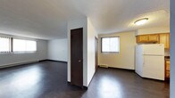 This single-floor space is characterized by its straightforward, functional layout and neutral design palette. The interior features dark hardwood-style flooring throughout, which contrasts with the white and light yellow walls to create a clean, understated aesthetic. A textured popcorn ceiling adds a classic touch overhead.

The layout is divided into distinct areas by a partial wall, creating a sense of separation between the open living zones without fully enclosing them. Dark-stained interior doors add a rich contrast against the lighter walls and contribute to the overall cohesive color scheme. Baseboards and dark trim details run consistently throughout the space, reinforcing the design continuity.

Natural light enters through a window fitted with vertical blinds, illuminating the adjacent area and providing a connection to the outdoors. A baseboard heating unit is positioned beneath the window, indicating a practical heating solution integrated into the space.

The open layout offers flexibility for furnishing and arrangement, making it adaptable for various uses. Overall, the space presents a clean, no-frills environment with functional features and a consistent, neutral design throughout.