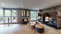 This thoughtfully designed single-floor amenity space blends productivity and comfort through a series of interconnected zones, all unified by warm herringbone-patterned wood flooring and a contemporary color palette of slate blue, grey, and natural wood tones. The layout, as revealed by the floor plan, follows a dynamic, winding configuration that encourages exploration while maintaining a cohesive aesthetic throughout.

The lounge area anchors the social heart of the space, featuring a sculptural sofa, organic ottoman clusters, and upholstered privacy pods with integrated lighting — ideal for quiet work or relaxed conversation. Large windows flood this zone with natural light and offer views of surrounding greenery, complemented by a textured reclaimed wood accent wall and curated abstract artwork.

Transitioning toward the work-focused area, a dedicated business center houses desktop computers at a sleek concrete-topped workstation, framed by industrial-style black metal and frosted glass partition doors. Adjacent cabinetry in charcoal grey pairs with a marble-look countertop, subway tile backsplash, illuminated glass shelving, and a matte black sink, lending the space a refined yet functional character. High-top bar seating along the window perimeter provides additional flexible workspace, completing a well-appointed environment designed for both collaboration and focused productivity.