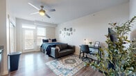 This thoughtfully designed studio unit offers a single-floor, open-concept layout that maximizes every square foot. Light-toned hardwood flooring runs throughout, creating a cohesive and airy feel across all areas of the space.

The combined living and sleeping area is bright and functional, featuring a ceiling fan with light fixture and a large window with balcony access that allows natural light to fill the room. A dedicated workspace with a desk and chair is neatly tucked alongside the sleeping area, while wall-mounted framed art adds a personal touch above.

The kitchen runs along one wall in a galley-style configuration, outfitted with stainless steel appliances including a refrigerator, electric range, microwave, and dishwasher. White upper and lower cabinetry with dark hardware provides ample storage, while granite-style countertops add a polished finish. The adjacent living area accommodates a sofa and television setup, making efficient use of the shared open space.

The bathroom is clean and modern, featuring a granite-topped vanity with under-sink storage, a large mirror, and a combination tub and shower with tiled surround. Consistent light wood flooring and neutral tones carry through from the main living space, maintaining a unified aesthetic throughout the unit.