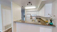 This single-floor residence offers a practical and well-organized layout designed for comfortable everyday living. The kitchen serves as a central highlight, featuring crisp white cabinetry that extends to the ceiling for ample storage, complemented by warm-toned countertops that add a subtle contrast to the bright, neutral color palette throughout. A wraparound breakfast bar defines the kitchen space, creating a natural division between the cooking area and the rest of the living space while also offering casual seating and additional counter space. White appliances, including a full-size refrigerator, electric range, built-in microwave, and a sink, are neatly integrated into the layout, contributing to the kitchen's clean and cohesive aesthetic. Recessed lighting and a track light fixture overhead ensure the space is well-lit, enhancing the open and airy feel. Smooth walls with a light, neutral finish run throughout, reflecting natural and artificial light effectively. A hallway visible from the kitchen hints at additional rooms beyond, suggesting a thoughtful floor plan that balances openness with private living areas. Overall, the space presents a tidy, functional, and welcoming environment.