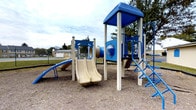 This outdoor playground is a well-equipped recreational space designed to encourage active play for children. The centerpiece is a multi-level play structure finished in blue and beige, featuring dual side-by-side slides that provide an inviting focal point. A horizontal tunnel connects sections of the structure, adding an element of exploration and adventure. On the right side, a blue metal ladder offers an alternative climbing route to the elevated platform, which is topped with a covered roof panel for partial shade. A separate blue curved slide extends to one side, providing additional play variety. The structure is supported by sturdy posts and incorporates a metallic sphere interactive element at its midsection. The ground surface beneath the equipment consists of packed gravel, designed to provide impact absorption. The surrounding area is open and grassy, offering ample space for free play. Mature deciduous trees line the perimeter, providing a natural backdrop and some shade. Residential buildings and a utility structure are visible in the background, suggesting this playground is situated within a neighborhood or community setting.