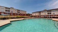 Located at 100 Patriot's Pointe Dr in Hillsborough, North Carolina, this outdoor amenity area centers around a generously sized swimming pool with clear, blue-green water and a distinctive curved shape. The pool is complemented by a separate shallow wading pool, making the space suitable for a range of swimmers. Surrounding the pool is a wide, well-maintained concrete sundeck furnished with multiple lounge chairs and patio seating shaded by blue market umbrellas, providing ample space for relaxation and outdoor enjoyment. A wooden pergola anchors one corner of the deck, offering a shaded gathering spot with additional seating. The area is enclosed by a white fence and accented with neatly maintained landscaping, including shrubs, ornamental grasses, and mature trees that provide natural shade and visual appeal. A brick utility or amenity building is positioned adjacent to the pool area, adding functional infrastructure to the space. The multi-story residential buildings surrounding the pool feature balconies that overlook the amenity area. The overall environment is open and bright, benefiting from abundant natural light and clear skies, creating an inviting outdoor retreat well-suited for warm-weather recreation and community gatherings.