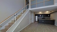 This two-story space showcases a thoughtfully designed open-concept layout that emphasizes vertical space and modern aesthetics. The main level features wood-look flooring that flows seamlessly throughout, anchoring the open living and kitchen areas. A striking staircase with glass panel railings and a clean white frame serves as a central architectural element, ascending to the upper level while allowing sightlines to travel freely through the space.

The kitchen is positioned toward the rear of the main floor and is outfitted with stainless steel appliances, including a full-size refrigerator and microwave. Dark cabinetry contrasts against a mosaic tile backsplash, and pendant lighting hangs above the kitchen island, which features a granite-style countertop. The upper level is visible from below through a windowed partition, contributing to the home's double-height ceiling effect and enhancing the sense of openness.

A louvered closet door beneath the staircase offers practical storage on the main floor. Neutral gray walls and recessed lighting throughout lend the space a clean, contemporary character, while the open-riser staircase design reinforces the modern, loft-inspired aesthetic.