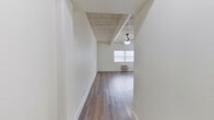 This 2-bedroom, 1-bathroom unit offers a practical and well-organized single-floor layout. Throughout the space, wood-look flooring provides a consistent and clean aesthetic, while crisp white walls contribute to a bright, open feel. A defining architectural feature is the exposed concrete beam ceiling, visible in the main living area and bedrooms, adding an industrial character to the otherwise minimalist design. The spacious primary bedroom is generously sized and includes a window for natural light and a wall-mounted air conditioning unit. The second bedroom also features a window with natural light and painted brick accent walls, adding texture to the room. The living area benefits from natural light through a large window, a ceiling fan, and a wall-mounted AC unit for climate control. The kitchen is efficiently designed with gray cabinetry, a granite-style countertop, a stainless steel sink, and an electric range. The bathroom is well-appointed with a full tub-shower combination, a white vanity with a mirrored medicine cabinet, and light wood-look tile flooring. Two hallways connect the rooms seamlessly, and a closet provides additional storage. Bold black interior doors with gold hardware add a stylish contrast throughout the unit.