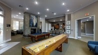 This single-floor amenity space is designed for recreation and social gatherings, offering a well-appointed layout that blends entertainment and functionality seamlessly. The game room serves as the centerpiece, featuring a classic billiards table with a blue felt surface and a full-length shuffleboard table, both finished in rich wood tones that complement the warm, neutral color palette of the space. A dramatic floor-to-ceiling stacked stone fireplace anchors one wall, serving as a striking architectural focal point and adding a cozy, upscale ambiance to the room. A large abstract artwork displayed above the fireplace adds a contemporary artistic touch. Recessed lighting throughout provides consistent, well-distributed illumination, while crown molding along the ceiling lends an air of refinement. Transitioning from the game area, a dining section offers seating for a group, positioned adjacent to a fully equipped kitchen. The kitchen features wooden cabinetry, stainless steel appliances including a refrigerator and microwave, and ample counter space, making it well-suited for hosting events. Framed botanical artwork decorates the walls near the kitchen, adding a finishing decorative detail. The carpeted flooring ties the entire space together with comfort and cohesion.
