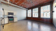 Located along the historic Euclid Avenue corridor in Cleveland, Ohio, this single-floor loft seamlessly blends industrial character with modern convenience. The open-concept main living area is defined by striking exposed brick walls and ceilings with original brick detailing, polished concrete floors that reflect natural light, and exposed ductwork that adds to the raw, urban aesthetic. A bank of large windows lines one wall, flooding the space with natural light while offering views of the surrounding neighborhood's classic brick architecture.

The kitchen is thoughtfully equipped with white raised-panel cabinetry, stainless steel appliances including a refrigerator, range, over-the-range microwave, and dishwasher, along with a double-basin sink and ample counter space. The layout creates a natural flow between the kitchen and the open living area, making the space highly functional.

The bathroom features tile flooring, a full tub-shower combination, a vanity with under-sink storage, and a large mirror with a dark-toned countertop shelf. Wall sconce lighting accents the living area, adding a warm contrast to the industrial elements overhead. Overall, this loft offers a distinctive urban living experience rooted in Cleveland's architectural heritage.