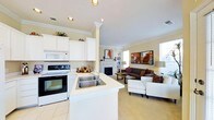 This single-story space showcases an open-concept layout that integrates the kitchen and living area into one cohesive, functional environment. The kitchen is outfitted with white cabinetry that extends to the upper walls, complemented by crown molding detailing that adds a classic, refined touch. A peninsula with a double stainless steel sink and light countertops serves as both a workspace and a natural divider between the kitchen and living zones. White appliances, including a built-in microwave and electric range, complete the kitchen setup.

The living area is carpeted in a neutral tone and centers around a fireplace with a white mantel, serving as a natural focal point. Recessed lighting throughout the space is supplemented by natural light streaming in through multiple windows. The room accommodates a comfortable seating arrangement with a sofa, armchairs, and a coffee table, creating an inviting atmosphere. Wall art and decorative accents add warmth and personality. Crown molding continues throughout, tying the kitchen and living areas together with consistent architectural detail.