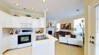 This single-story space showcases an open-concept layout that integrates the kitchen and living area into one cohesive, functional environment. The kitchen is outfitted with white cabinetry that extends to the upper walls, complemented by crown molding detailing that adds a classic, refined touch. A peninsula with a double stainless steel sink and light countertops serves as both a workspace and a natural divider between the kitchen and living zones. White appliances, including a built-in microwave and electric range, complete the kitchen setup.

The living area is carpeted in a neutral tone and centers around a fireplace with a white mantel, serving as a natural focal point. Recessed lighting throughout the space is supplemented by natural light streaming in through multiple windows. The room accommodates a comfortable seating arrangement with a sofa, armchairs, and a coffee table, creating an inviting atmosphere. Wall art and decorative accents add warmth and personality. Crown molding continues throughout, tying the kitchen and living areas together with consistent architectural detail.