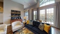 Located at 27 Station Ct in the heart of Greenville, SC, this sophisticated single-floor space offers a thoughtfully designed interior that blends architectural elegance with warm, contemporary style. The grand entryway sets a striking first impression, featuring soaring vaulted ceilings with a cathedral glass transom window, rich dark hardwood floors, and a statement chandelier. A built-in bench with colorful accent pillows and a sleek indoor water feature flank the entry, creating a welcoming and refined arrival experience. Large French doors flood the space with natural light and provide direct views to the outdoors. The main lounge area continues the refined aesthetic with expansive floor-to-ceiling windows and French doors that frame outdoor greenery, complemented by dramatic high ceilings with clerestory glazing above. The sitting area is furnished with a deep navy sofa accented by yellow and patterned throw pillows, anchored by a vibrant area rug. A charming "McBee Station" chalkboard display with a coffee and beverage bar adds character and a community-oriented touch. Throughout, neutral warm-gray walls, tall baseboards, recessed lighting, and consistent hardwood flooring create a cohesive, polished atmosphere that is both inviting and functional.