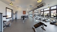 This fitness center offers a comprehensive and thoughtfully designed workout environment across a single, expansive floor. The main exercise area is generously proportioned, accommodating a wide array of cardio equipment including treadmills, elliptical trainers, and rowing machines, all positioned along a wall of large, multi-pane windows that flood the space with natural light and provide views of the surrounding outdoors. A dedicated free weights section features a full dumbbell rack and adjustable benches, complemented by a variety of strength training machines for a complete full-body workout. Exposed industrial-style ductwork runs along the ceiling, lending the space a modern, urban aesthetic, while stylish pendant lights provide warm, focused illumination throughout. Durable rubber flooring covers the entire workout area, ensuring both safety and comfort. A separate cycling studio houses multiple stationary bikes and is accessible through glass-paneled doors, offering privacy for group or individual sessions. An additional mirrored studio space with yoga mats and stability balls provides a dedicated area for stretching, yoga, or functional training. Motivational artwork adorns the white walls, contributing to an energetic and welcoming atmosphere. The overall design balances functionality with a clean, modern aesthetic.