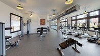 This fitness center offers a comprehensive and thoughtfully designed workout environment across a single, expansive floor. The main exercise area is generously proportioned, accommodating a wide array of cardio equipment including treadmills, elliptical trainers, and rowing machines, all positioned along a wall of large, multi-pane windows that flood the space with natural light and provide views of the surrounding outdoors. A dedicated free weights section features a full dumbbell rack and adjustable benches, complemented by a variety of strength training machines for a complete full-body workout. Exposed industrial-style ductwork runs along the ceiling, lending the space a modern, urban aesthetic, while stylish pendant lights provide warm, focused illumination throughout. Durable rubber flooring covers the entire workout area, ensuring both safety and comfort. A separate cycling studio houses multiple stationary bikes and is accessible through glass-paneled doors, offering privacy for group or individual sessions. An additional mirrored studio space with yoga mats and stability balls provides a dedicated area for stretching, yoga, or functional training. Motivational artwork adorns the white walls, contributing to an energetic and welcoming atmosphere. The overall design balances functionality with a clean, modern aesthetic.