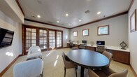 This single-floor space functions as a well-appointed business center and lounge, thoughtfully designed to accommodate both individual productivity and group collaboration. The room is finished with neutral-toned walls, rich dark wood crown molding and baseboards, and a light herringbone-patterned carpet that lends a clean, professional aesthetic throughout.

A built-in workstation runs along one wall, outfitted with multiple desktop computers, ergonomic task chairs, a printer, and warm wood cabinetry below, providing ample workspace for several users simultaneously. A row of framed windows above the workstation admits natural light while maintaining a focused atmosphere. Centrally positioned in the room, a round dark wood table surrounded by upholstered chairs serves as an ideal meeting or collaborative work area.

A dedicated lounge zone offers white accent chairs paired with a leather ottoman and a wall-mounted flat-screen television, creating a comfortable setting for informal gatherings or presentations. Framed artwork accents the walls, adding a polished touch.

Prominent dark wood French doors with glass panes divide the space and connect to an adjacent outdoor or interior area, allowing natural light to filter through and enhancing the overall sense of openness. Recessed ceiling lighting ensures consistent, even illumination throughout the room.