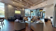 Located at 1825 N Las Palmas Ave in the heart of Hollywood, this single-floor communal space combines contemporary architecture with a curated artistic sensibility. The expansive open-plan layout is anchored by rich hardwood flooring, soaring double-height ceilings, and an impressive wall of floor-to-ceiling black-framed glass that floods the interior with natural light and offers views of the surrounding neighborhood.

The main lounge area features a refined mix of patterned armchairs and curved sofas arranged around glass and gold coffee tables, accented by an array of globe and drum pendant lighting overhead. A dramatic stone accent wall serves as the backdrop for a linear fireplace and mounted display screen, adding warmth and texture to the space. Curated artwork is displayed throughout, including a bold, large-scale contemporary painting and framed fine art pieces, lending the space a gallery-like quality.

Multiple distinct seating zones cater to varied uses, from intimate conversation nooks with cream channel-tufted banquette seating and black marble cube tables, to casual dining areas with modern chairs and wooden tables positioned beside exterior access points. Built-in cabinetry with open shelving displays sculptural objects, reinforcing the space's attention to artistic detail. Overall, the environment projects a polished, upscale ambiance ideal for socializing and relaxation.