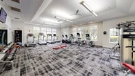 Located at 4122 Avalon Dr in Peabody, Massachusetts, this fitness center offers a comprehensive and thoughtfully designed exercise space across a single floor. The room is generously proportioned, providing ample space for a wide variety of workouts without feeling crowded.

The interior design is clean and contemporary, featuring crisp white walls accented with subtle textured tile, tray ceilings with recessed lighting, and ceiling fans that promote air circulation throughout. A bold, patterned carpet in shades of grey and black covers the floor, lending a modern aesthetic while providing cushioning underfoot.

Natural light pours in through large, multi-pane windows positioned along multiple walls, offering pleasant views of surrounding greenery and creating a bright, energizing atmosphere. A glass-paneled door provides direct access to the outdoors.

The fitness center is well-stocked with a diverse range of equipment, including multiple treadmills, elliptical trainers, stationary and recumbent bikes, a cable strength machine, and a dedicated free weights and medicine ball station. A flat-screen television is mounted on the wall, and mirrored surfaces help enhance the sense of openness. A small amenity station is also present, adding a convenient touch to the overall experience. The space strikes an effective balance between functionality and inviting design.