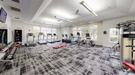 Located at 4122 Avalon Dr in Peabody, Massachusetts, this fitness center offers a comprehensive and thoughtfully designed exercise space across a single floor. The room is generously proportioned, providing ample space for a wide variety of workouts without feeling crowded.

The interior design is clean and contemporary, featuring crisp white walls accented with subtle textured tile, tray ceilings with recessed lighting, and ceiling fans that promote air circulation throughout. A bold, patterned carpet in shades of grey and black covers the floor, lending a modern aesthetic while providing cushioning underfoot.

Natural light pours in through large, multi-pane windows positioned along multiple walls, offering pleasant views of surrounding greenery and creating a bright, energizing atmosphere. A glass-paneled door provides direct access to the outdoors.

The fitness center is well-stocked with a diverse range of equipment, including multiple treadmills, elliptical trainers, stationary and recumbent bikes, a cable strength machine, and a dedicated free weights and medicine ball station. A flat-screen television is mounted on the wall, and mirrored surfaces help enhance the sense of openness. A small amenity station is also present, adding a convenient touch to the overall experience. The space strikes an effective balance between functionality and inviting design.