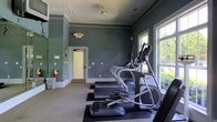 This fitness center is a single-floor facility designed to support an active lifestyle in a comfortable and well-appointed environment. The space is finished with sage-green walls accented by crisp white crown molding and baseboards, giving it a polished, classic aesthetic that sets it apart from a typical gym setting.

Large windows along one wall flood the interior with abundant natural light, offering pleasant outdoor views of surrounding greenery and creating an open, airy atmosphere during workouts. The ceiling features recessed lighting supplemented by a ceiling fan, ensuring the space remains well-lit and comfortable.

The fitness center is equipped with a row of cardio machines, including treadmills and elliptical trainers, positioned to take full advantage of the natural light from the windows. Rubber-matted flooring beneath the equipment protects the carpeted surface and provides stability during use.

Safety and accessibility are clearly prioritized, with a prominently mounted fire extinguisher and an illuminated exit sign near the entry door. The entryway features a classic paneled door with an adjacent staircase visible beyond, providing straightforward access to the facility. Overall, this fitness center combines functional equipment with refined architectural details to create an inviting and practical workout environment.