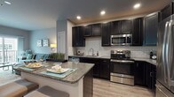 This single-floor residence offers a thoughtfully designed layout that balances modern aesthetics with everyday functionality. The kitchen is a standout feature, showcasing dark espresso cabinetry, a classic white subway tile backsplash, granite countertops, and a full suite of stainless steel appliances including a French door refrigerator, range, over-the-range microwave, and dishwasher. A peninsula island with bar seating provides an ideal space for casual dining and entertaining, flowing seamlessly into the rest of the unit. The living area is bright and welcoming, with wide-plank wood-style flooring, neutral gray walls, and sliding glass doors that open onto a private balcony, allowing for natural light and outdoor views. The bedroom is generously sized, featuring plush carpeting, a large window that fills the room with light, and a stylish sliding barn door that leads to a spacious walk-in closet with built-in organization. The bathroom is finished with large-format tile flooring, a dark vanity, and a full shower-tub combination. Throughout the space, recessed lighting and a cohesive neutral color palette create a modern, comfortable ambiance.