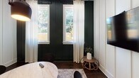 This thoughtfully designed single-floor space makes a striking impression through its bold use of contrasting colors and carefully curated finishes. The main living area showcases crisp white board-and-batten wall paneling that stretches from floor to ceiling, creating a clean, structured backdrop that highlights the rich warmth of the dark hardwood floors beneath. A wall-mounted flat-screen television anchors one side of the room, while a geometric wood credenza with chevron detailing and rose gold accents serves as a stylish focal point on the opposite wall, topped with a framed mirror and contemporary lamp. A round marble-top table at the center of the room is paired with sleek black accent chairs, all resting atop a woven area rug that adds texture and warmth to the space. One of the most dramatic design elements is the deep forest green board-and-batten accent wall, which provides a rich contrast to the white paneling and frames two tall windows dressed with sheer white curtains. These windows invite generous natural light and offer views of lush outdoor greenery. An elegant arched doorway with dark wood trim leads to an adjacent green-accented room, adding architectural character and visual depth. A modern pendant light hangs above the central table, completing the layered, eclectic yet cohesive aesthetic of this well-appointed space.