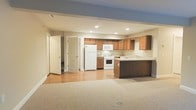 This single-floor space offers an open-concept design that seamlessly connects the living area to the kitchen. The living space is finished with neutral-toned carpet, providing a comfortable foundation, while the kitchen area transitions to smooth hardwood-style flooring. Recessed lighting throughout the ceiling ensures consistent, well-distributed illumination across the entire space.

The kitchen is a functional highlight, featuring warm medium-toned wood cabinetry arranged along two walls in an L-shaped configuration. A dark-finished kitchen island provides additional counter space and storage. White appliances — including a refrigerator, range, built-in microwave, and a sink set into light-colored countertops — complete the kitchen setup. A tile backsplash adds a clean, finished look behind the appliances.

Two white panel doors are visible along the adjacent wall, providing access to additional rooms or storage areas. The neutral beige and warm-toned color palette throughout creates a cohesive, understated ambiance. The layout is practical and well-organized, making efficient use of the available space.