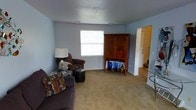 This two-floor home offers a practical and comfortable layout designed for everyday living. The main living area is finished with wall-to-wall carpet in a warm neutral tone, creating a soft and inviting atmosphere underfoot. Light blue-grey walls lend the space a calm, understated character, while a textured ceiling adds subtle visual depth overhead.

A window positioned along the far wall allows natural light to filter into the room, brightening the interior and contributing to an open feel. The living area provides ample space for seating arrangements and furnishings, as evidenced by the room's capacity to accommodate a sofa, armchair, ottoman, and a substantial wooden armoire with decorative cabinetry.

A doorway visible along the side wall leads to adjacent areas of the home, where hardwood or laminate flooring can be seen transitioning into what appears to be an entryway or hallway. Stairway access is also visible, connecting this main level to the second floor of the home.

The overall ambiance is relaxed and functional, with a straightforward design that emphasizes usability. The neutral color palette throughout provides a versatile backdrop suited to a variety of décor styles.