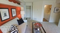 This single-floor space makes a strong visual impression through its thoughtful use of color and layout. The living area serves as the central gathering space, anchored by a striking burnt-orange accent wall that adds warmth and personality against the surrounding neutral beige tones. Built-in shelving above provides a display ledge, adding architectural interest to the room.

The living area is carpeted and comfortably sized, accommodating a tufted sofa, accent chairs, a glass coffee table, and side tables with ease. A closet with bifold doors opens directly off the living space, offering convenient storage. Floor vents suggest a forced-air climate system throughout.

Natural transitions lead from the living area into adjacent spaces, including what appears to be a hallway connecting to additional rooms. A glimpse into the background reveals hard flooring in the adjoining area, providing a contrast to the soft carpet underfoot in the main living space.

Overall, the space presents a clean, functional layout with a warm, inviting ambiance. The combination of neutral walls, accent color, and practical storage solutions creates a well-balanced environment suitable for everyday living.