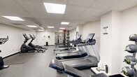 Located at 2920 5th Ave in Troy, New York, this fitness center offers a well-organized, single-floor exercise space designed to accommodate a full range of workout routines. The room features a drop-panel ceiling outfitted with recessed fluorescent lighting panels that provide bright, even illumination throughout. The flooring consists of durable speckled rubber, ideal for high-traffic gym use and equipment placement.

The cardio section is equipped with multiple treadmills from Spirit and Endurance brands, along with elliptical trainers from Sole and a stationary upright bike, providing ample options for cardiovascular workouts. Rowing machines are also present, adding variety to the cardio offerings. The strength training area features a multi-station cable machine system and a squat/power rack with free weights, supporting a comprehensive resistance training regimen. A TKO adjustable workout bench is available for free weight exercises.

A dedicated area in the rear of the space includes a wood-toned flooring section near the cable station, adding a slight design contrast to the otherwise uniform gray floor. A restroom is accessible directly within the space for user convenience. The clean, white walls and structured layout create a functional and focused environment, making this fitness center a practical and fully equipped facility for daily exercise.