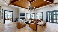 Located at 8585 Woodway Dr in Houston, TX, this single-floor clubhouse space offers a well-appointed and thoughtfully designed communal environment. The main lounge area immediately impresses with its soaring ceilings adorned with warm wood beam detailing and a striking wrought-iron chandelier that anchors the room. Natural flagstone flooring flows throughout, lending an earthy, upscale character to the space. A textured accent wall frames a sleek linear fireplace topped by a large flat-screen display, creating an inviting focal point. Generous seating, including leather lounge chairs and a plush sofa, is arranged to encourage conversation and relaxation. Built-in shelving units provide both decorative and functional storage. An abundance of natural light pours in through large sliding glass doors and floor-to-ceiling windows with bold navy blue trim, offering direct views and access to an outdoor patio and pool area. A dedicated dining nook with café-style tables and chairs sits adjacent to the glass-lined exterior wall. The kitchen is efficiently laid out with granite countertops, ample cabinetry, a range, and a coffee station, making it ideal for community gatherings. A quieter seating area near windowed office nooks provides a more subdued setting for focused activities.