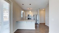 Located at 2170 Snyder Circle in the desirable Mount Pleasant, South Carolina community, this single-floor residence offers a clean, contemporary design throughout. The open-concept kitchen serves as a central highlight of the home, showcasing light-toned cabinetry that extends to the ceiling for ample storage, a classic subway tile backsplash, and a full suite of stainless steel appliances including a refrigerator, range, and built-in microwave. A peninsula with a granite-style countertop provides additional prep space and casual seating, making it ideal for entertaining or everyday living. Elegant pendant lighting above the peninsula, paired with a modern track light fixture, creates a warm and well-lit atmosphere. Rich, dark hardwood-style flooring runs throughout, offering a striking contrast to the bright white cabinetry and light walls. The open layout connects the kitchen seamlessly to the adjacent living area, enhancing the sense of space and flow. Natural light filters in through windows fitted with blinds, adding to the airy, inviting ambiance of this well-appointed single-story home.