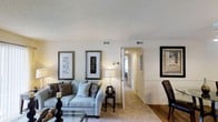 Located at 5650 Netherfield in Fayetteville, North Carolina, this single-floor residence offers a thoughtfully designed layout with a warm and inviting ambiance throughout. The living area serves as the heart of the home, featuring soft, neutral wall tones and plush wall-to-wall carpeting that creates a cozy and comfortable atmosphere. A textured ceiling adds subtle architectural character to the space, while multiple ceiling vents ensure consistent climate control throughout. The living room provides ample space for relaxed seating arrangements, with natural light complemented by warm lamp lighting that enhances the welcoming feel of the room. A wide doorway connects the living area to a hallway that leads further into the home, suggesting a well-organized and functional floor plan. The transition between spaces is seamless, with consistent flooring and neutral color palettes that lend a cohesive aesthetic throughout. Classic chair rail detailing along the walls adds a touch of traditional elegance to the dining area, which flows naturally from the main living space. Overall, this home presents a clean, move-in-ready interior with timeless design choices that offer a comfortable and practical living environment in a residential Fayetteville neighborhood.