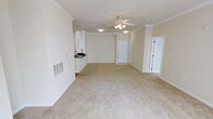 This single-floor residence offers an open and airy layout that seamlessly connects the main living area to the kitchen. The expansive living space is finished with plush, neutral-toned carpeting that lends a clean and comfortable feel underfoot. A ceiling fan with integrated lighting is centrally positioned, ensuring both airflow and illumination throughout the room.

Crown molding runs along the perimeter of the ceilings, adding a refined architectural detail that elevates the overall aesthetic. Decorative columns subtly define the transition between spaces, contributing to the home's sense of structure without enclosing the open floor plan.

The kitchen is partially visible from the main living area and features white cabinetry, a dark countertop, and a sink — offering a practical and tidy workspace. The crisp white walls and trim throughout the space reflect natural and artificial light effectively, creating a bright and welcoming ambiance.

Two interior doors are visible, suggesting additional private rooms or utility spaces beyond the main living area. A wall-mounted HVAC vent contributes to year-round climate control. Overall, the space presents a well-maintained, functional, and inviting environment with clean finishes and a neutral palette that offers flexibility for a variety of furnishing styles.