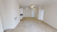 This single-floor residence offers an open and airy layout that seamlessly connects the main living area to the kitchen. The expansive living space is finished with plush, neutral-toned carpeting that lends a clean and comfortable feel underfoot. A ceiling fan with integrated lighting is centrally positioned, ensuring both airflow and illumination throughout the room.

Crown molding runs along the perimeter of the ceilings, adding a refined architectural detail that elevates the overall aesthetic. Decorative columns subtly define the transition between spaces, contributing to the home's sense of structure without enclosing the open floor plan.

The kitchen is partially visible from the main living area and features white cabinetry, a dark countertop, and a sink — offering a practical and tidy workspace. The crisp white walls and trim throughout the space reflect natural and artificial light effectively, creating a bright and welcoming ambiance.

Two interior doors are visible, suggesting additional private rooms or utility spaces beyond the main living area. A wall-mounted HVAC vent contributes to year-round climate control. Overall, the space presents a well-maintained, functional, and inviting environment with clean finishes and a neutral palette that offers flexibility for a variety of furnishing styles.