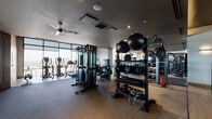 This well-appointed fitness center in Denver's Cherry Creek neighborhood offers a comprehensive workout environment designed with both functionality and aesthetics in mind. The space features a sophisticated dark ceiling with recessed lighting and ceiling fans, contrasted by warm wood-paneled accent walls that lend a refined, contemporary character throughout.

The cardio zone takes full advantage of floor-to-ceiling windows, providing panoramic views of the surrounding Denver skyline while using ellipticals, treadmills, and stair climbers. Natural light floods this area, creating an energizing atmosphere for cardio training.

The strength training area is extensively equipped with a multi-station cable machine, a full rack of Matrix and Torque brand equipment, adjustable benches, and a large selection of dumbbells organized on tiered racks. Medicine balls, kettlebells, and resistance accessories are neatly stored for functional training needs.

A dedicated free weights and functional training zone features rubber flooring for safety and durability, a large-format wall-mounted display screen, and expansive floor-to-ceiling mirrors that visually enlarge the space. The entry area showcases warm hardwood flooring and framed glass double doors, providing a polished transition into the facility. Overall, the space delivers a premium, hotel-caliber fitness experience.