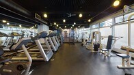This single-floor fitness center on Ocean Avenue in Santa Monica offers a comprehensive workout facility with a thoughtfully arranged layout. The space is divided into distinct zones, including a dedicated cardio area lined with multiple treadmills and additional aerobic machines positioned along a mirrored wall, which enhances the sense of openness throughout the floor. Large windows along one side of the facility allow natural light to filter in, providing views of the surrounding streetscape.

The strength training section features a wide selection of cable and weight machines arranged to maximize usable floor space, all set atop durable dark rubber flooring. Pendant globe lights hang from the dramatic black ceiling, which also exposes industrial ductwork, lending the space a modern, urban aesthetic.

A particularly striking architectural feature is a cylindrical structure finished in bold blue-and-white horizontal stripes, fitted with a distressed metal door with porthole windows — a clear nod to Santa Monica's coastal character. Adjacent to this feature wall is a free weights and functional training area stocked with dumbbells, stability balls, and benches. The overall ambiance is energetic and purposefully designed, balancing industrial styling with a beach-inspired personality.