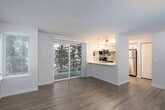 This single-floor residence offers a thoughtfully designed layout that balances comfort and functionality. The main living area is bright and welcoming, featuring durable hardwood-style flooring, neutral-toned walls, and a corner window arrangement that invites abundant natural light. A sliding glass door opens to a private balcony with leafy tree views, creating a seamless indoor-outdoor connection and a serene atmosphere.

The kitchen is positioned adjacent to the living area via a peninsula-style breakfast bar, making it ideal for both everyday use and casual entertaining. It is outfitted with crisp white cabinetry, a stylish mosaic tile backsplash, light countertops, and a full suite of stainless steel appliances including a range, over-the-range microwave, and refrigerator. Small windows above the counters bring in additional natural light.

The bedroom is a quiet, carpeted retreat with clean walls and ample outlet placement, offering a calm and restful environment. A second room with hardwood-style flooring provides flexible use as an additional bedroom or living space. The bathroom features a wide vanity with a single-basin sink, generous mirror, and clean white finishes throughout, maintaining the residence's consistent, polished aesthetic.
