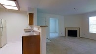 This single-floor home offers an open and functional layout designed for comfortable everyday living. The kitchen features warm wood cabinetry, a granite-style countertop, and a built-in sink, all framed by a half-wall pass-through that connects seamlessly to the main living area. This design choice promotes an open feel while maintaining a defined cooking space. An upper cabinet with a mirrored backing adds a subtle decorative touch to the kitchen zone.

The living area is finished with wall-to-wall carpet that lends a warm and quiet atmosphere to the space. A wood-trimmed fireplace with a classic tile surround serves as a natural focal point, adding character and a cozy ambiance to the room. The neutral wall tones throughout create a clean, versatile backdrop suitable for a variety of interior styles.

A doorway from the main living area leads into an additional carpeted room, offering flexibility for use as a bedroom, office, or den. The space is bathed in soft, even lighting, contributing to a calm and inviting environment. Overall, this home presents a straightforward and practical layout with thoughtful architectural details that balance function and comfort throughout.