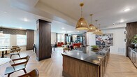 Located in Smyrna, Georgia, this thoughtfully designed single-floor community amenity space seamlessly blends social gathering and relaxed comfort into one cohesive open-plan layout. The herringbone-patterned hardwood flooring flows throughout, providing a warm and sophisticated foundation that unifies each distinct zone.

The kitchen area serves as a central focal point, anchored by an expansive dark granite island with bar seating. Rich walnut cabinetry lines the walls beneath a lustrous tile backsplash, while a trio of woven rattan pendant lights suspended on brass chains add warmth and texture overhead. The kitchen is fully equipped with a deep sink, generous counter space, and ample storage.

Adjacent to the kitchen, a lounge area invites relaxed gathering, furnished with vibrant red and coral upholstered sofas, a wood coffee table, and glass-fronted display bookcases framed in dark metal. A sculptural brass-and-black linear chandelier illuminates the space, while large windows with layered drapery allow natural light to fill the room.

A café-style banquette area completes the layout, featuring plush booth seating along a dramatic navy botanical mural wall, marble-topped bistro tables, and curated framed artwork. Natural light enters through multiple windows, enhancing the inviting ambiance. Together, these zones create a versatile, design-forward communal space ideal for socializing and everyday gatherings.
