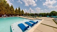 The outdoor pool area at The Luxe of Southaven in Southaven, Mississippi, offers a well-appointed recreational space designed for comfort and leisure. The centerpiece is a generously sized swimming pool with clear, blue-green water, bordered by a broad concrete sundeck that provides ample room for relaxation and circulation. A distinctive feature of the pool is the inclusion of submerged loungers positioned directly in the shallow water, allowing residents to recline while remaining partially immersed—an amenity that elevates the overall resort-style experience.

The sundeck is lined with teal-cushioned chaise loungers arranged along multiple sides of the pool, offering abundant seating options in both shaded and sun-exposed areas. Patches of artificial turf accent the deck, adding visual contrast and a softer underfoot surface in select zones. The perimeter of the pool area is enclosed by a dark metal fence, providing security while maintaining visibility to the surrounding greenery. Mature trees and dense shrubs frame the space, contributing natural privacy and shade. An adjacent brick building with multiple entry doors serves as a facility structure supporting the pool area. The overall ambiance is open, airy, and well-suited for warm-weather outdoor activity.