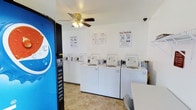 This single-floor laundry facility is designed for straightforward, self-service use. The main area houses a row of top-loading washing machines lined against a clean white wall, each equipped with coin-operated payment systems. Instructional posters, including "Easy Washing" and "Simple Drying" guides, are mounted on the wall to assist users through the laundry process. A beverage vending machine is conveniently positioned near the entrance, adding a practical amenity for patrons. The room features light tile flooring that is easy to maintain, and a ceiling fan with an integrated light fixture provides both ventilation and ambient lighting. A security camera is mounted near the ceiling, ensuring the safety of the space. The overall environment is clean, functional, and well-organized, with a no-frills layout focused on efficiency. A small table or counter is partially visible, likely available for folding or sorting laundry. The white walls and bright lighting contribute to a clean, utilitarian atmosphere suited for its purpose.