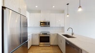 This single-floor residence in Weymouth, MA offers a thoughtfully designed layout with modern finishes throughout. The open-concept kitchen is a standout feature, showcasing two-tone cabinetry with light wood lower cabinets and high-gloss white upper cabinets extending to the ceiling for ample storage. A classic white subway tile backsplash complements the stainless steel appliances, including a range, over-the-range microwave, refrigerator, and dishwasher. Light quartz countertops and an undermount sink complete the space, while pendant lighting adds a warm, contemporary touch. The kitchen flows seamlessly into the living area, which is bright and open, finished with warm wood-look flooring that continues throughout the unit. The bedroom features a large black-framed window that floods the room with natural light and offers views of the surrounding treeline. Crisp white walls and clean lines maintain a consistent modern aesthetic. The bathroom is finished with large-format white tile in the tub/shower surround, a light wood vanity with a white quartz countertop, and polished chrome fixtures, creating a clean, spa-like feel. Recessed lighting throughout enhances the overall brightness of the space.