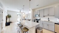 Located at 3755 E McKinney St in Denton, TX, this well-appointed one-bedroom, one-bathroom residence offers a thoughtfully designed open-concept layout with modern finishes throughout. The kitchen and living area flow seamlessly together, anchored by a large granite-topped island with bar seating illuminated by stylish pendant lighting. The kitchen is equipped with gray shaker-style cabinetry, a full suite of stainless steel appliances including a range, microwave, dishwasher, and refrigerator, and a white subway tile backsplash. Light hardwood-style flooring runs throughout the main living areas, enhancing the bright and airy atmosphere. The living room receives natural light through a glass exterior door with a transom window and is finished with a ceiling fan overhead. The bedroom features plush carpet flooring, a ceiling fan, and a window with outdoor views, providing a calm and comfortable retreat. A dedicated walk-in closet offers generous built-in shelving and dual hanging rods for ample storage. The bathroom continues the refined aesthetic with gray cabinetry, granite countertops, black hardware accents, and light wood-style flooring. The overall design is clean, contemporary, and functional throughout.